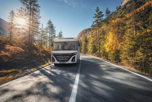 Road in autumn forest at sunset in Italy. Beautiful mountain roadway, trees with orange foliage and sunlight. Landscape with empty asphalt road through woodland, blue sky in fall. Transportation